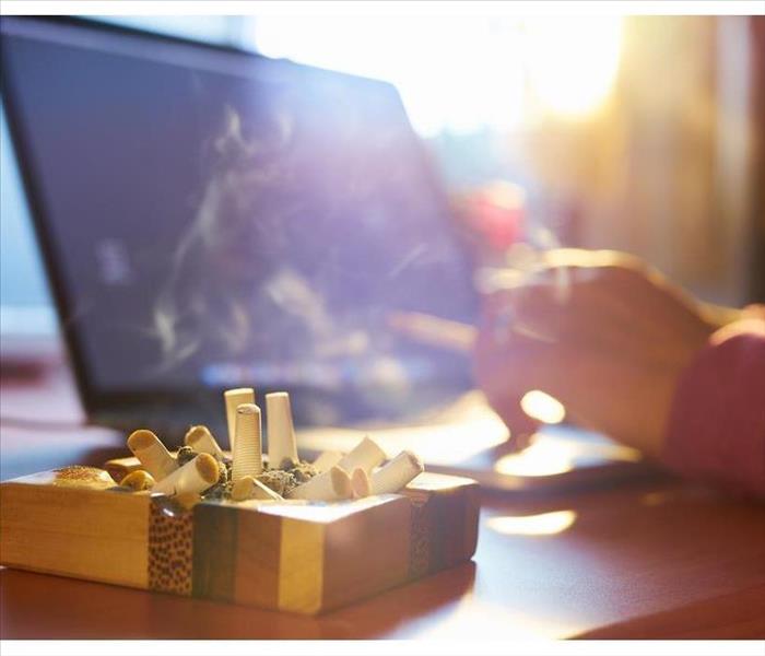Close up of ashtray full of cigarette, with man in background working on laptop computer and smoking indoors on early morning